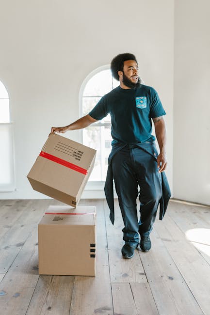 A man with curly hair and a beard, dressed in a blue t-shirt and dark trousers, is sitting on a wooden floor inside a room with two large arched windows allowing natural light to illuminate the space.