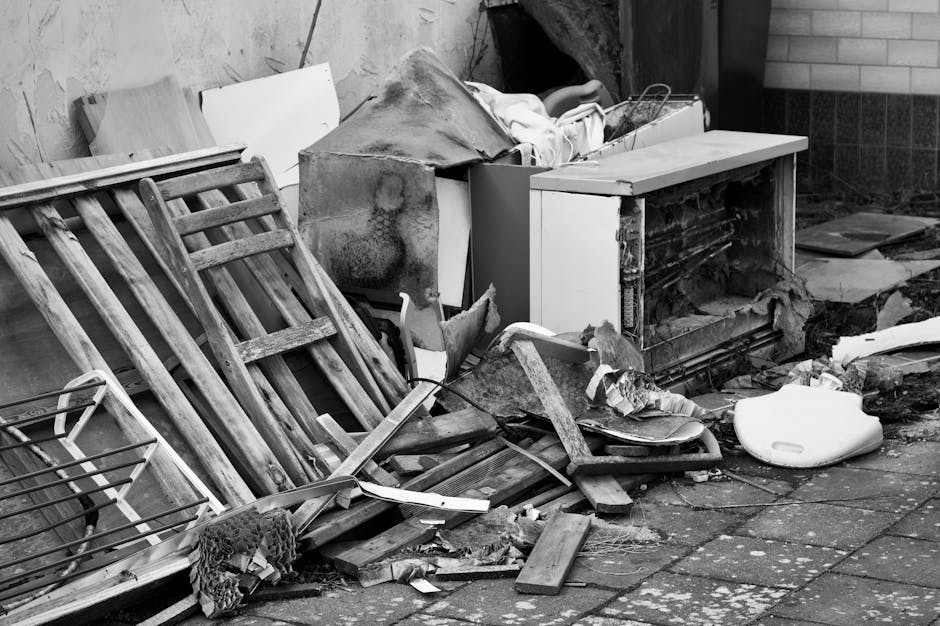A cluttered outdoor area with discarded household waste and debris, including a variety of wooden furniture such as a dismantled bed frame and a small table, alongside broken pieces of wood, cardboard