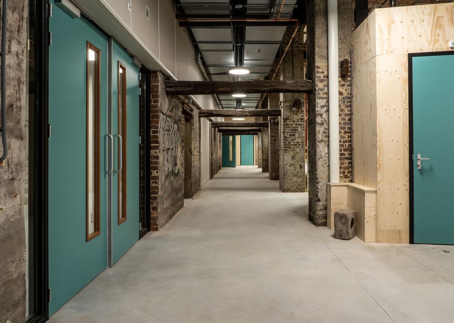 A long corridor inside an industrial warehouse space, featuring exposed brick walls and wooden beams supporting the ceiling. On the left side, there are large, teal-colored metal sliding doors with ve