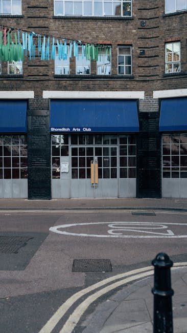 A three-story rounded building with a historic appearance located at a busy street corner, featuring a black facade on the ground level with gold accents and signage for the Old Blue Last. The upper f