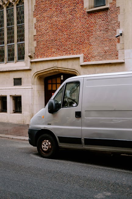 A vintage green camper van with a grey roof parked on a city street in Shoreditch, displaying the side profile of the vehicle. The van features a large window with horizontal metal bars for ventilatio
